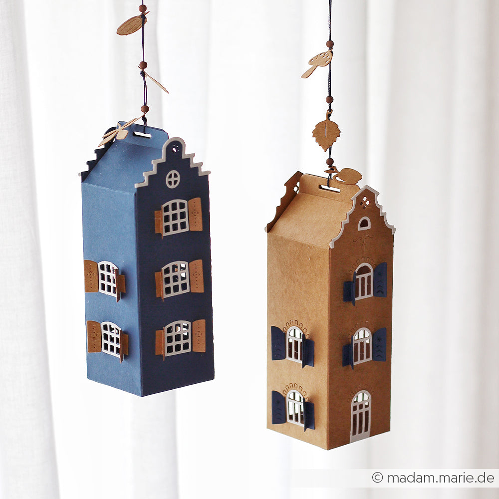 Zwei hängende Papierlaternen in Form von hohen Häusern, eine blau und eine braun, mit ausgeschnittenen Fenstern und Türen. Diese bezaubernden Laternen wurden aus Designpapieren und mit der Motivstanze „Häuserbox 4“ von DieCut Systeme gefertigt und verleihen jedem Raum eine verspielte Note.