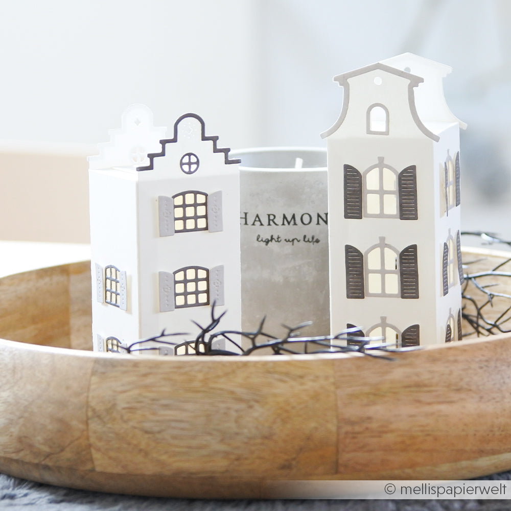 Papierlaternen, die aus eleganten Designpapieren gefertigt und mit der Motivstanze „Häuserbox 3“ von DieCut Systeme ausgestanzt wurden, stehen mit einer Kerze in einer Holzschale neben dekorativen schwarzen Zweigen.