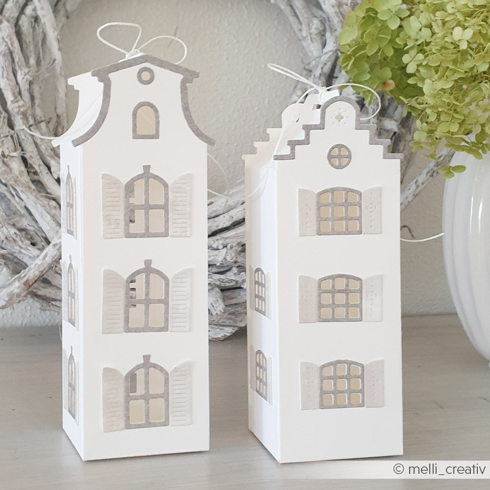 Zwei weiße, dekorative Papierhäuschen aus hochwertigem Designpapier mit Fenstern und grauen Akzenten stehen auf einem Tisch neben grünen Hortensien. Die bezaubernden Häuschen wurden mit der Motivstanze „Häuserbox 3“ ausgestanzt, um präzise Details zu erzielen.