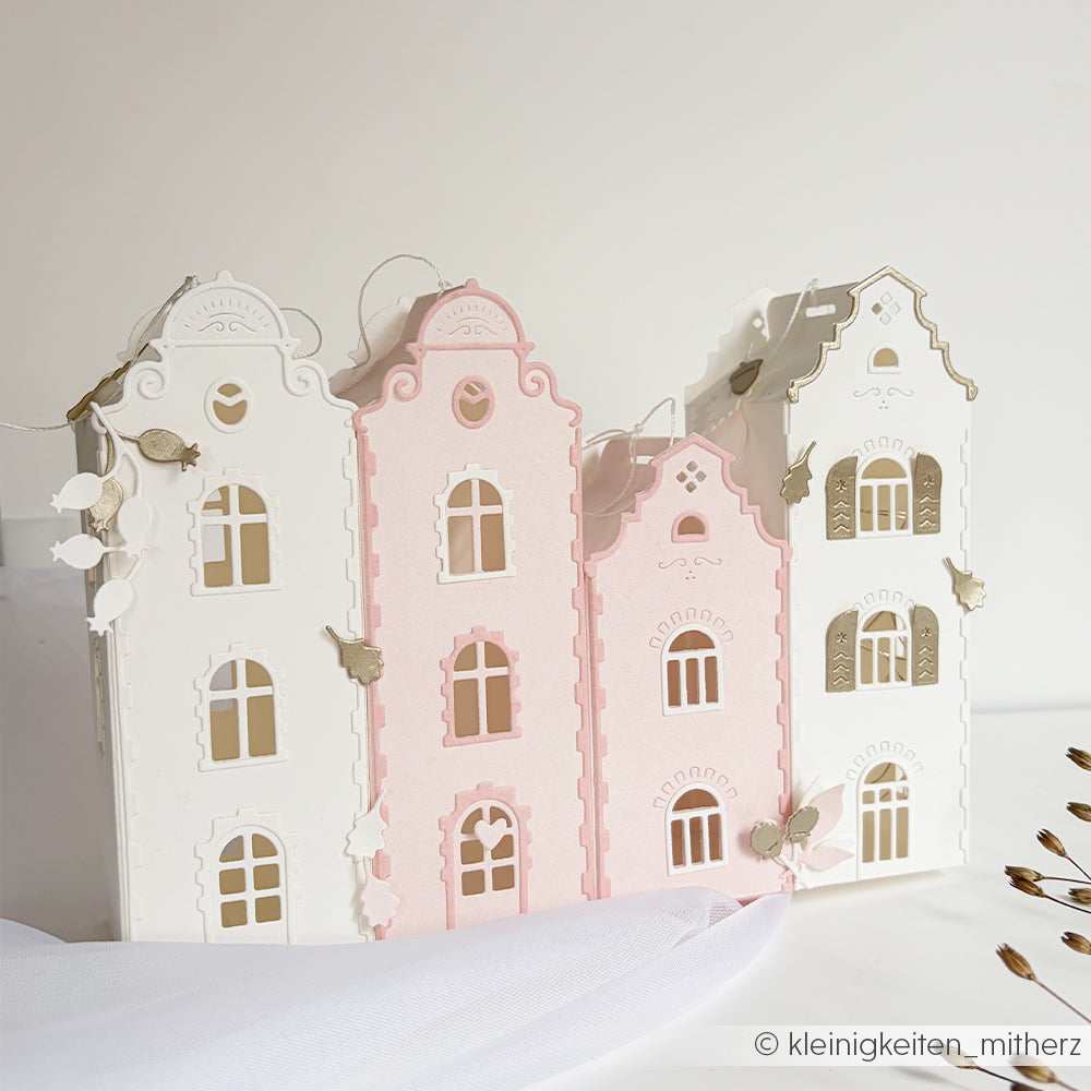 Drei pastellfarbene Papierhäuser in Weiß und Rosa mit goldenen Fenstern, gefertigt aus erlesenen Designpapieren mit der Motivstanze „Häuserbox 4 Stanzsysteme“, elegant präsentiert auf einem weißen Untergrund.