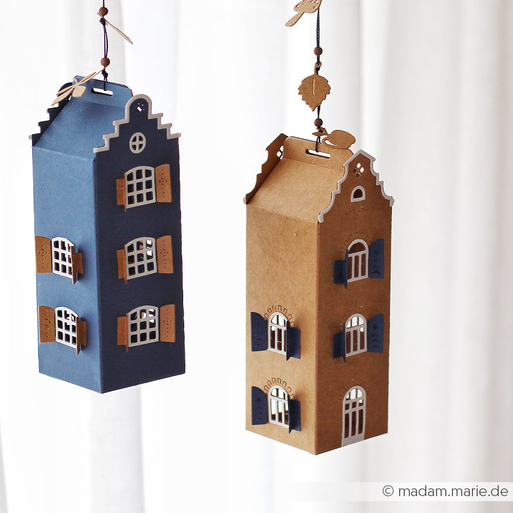Zwei hängende Papierlaternen in Form von hohen Häusern mit Fenstern und Fensterläden, eine in Blau und eine in Braun, gefertigt aus edlen Designpapieren mit Hilfe des Stanzsystems „Motivstanze Häuserbox 3“.