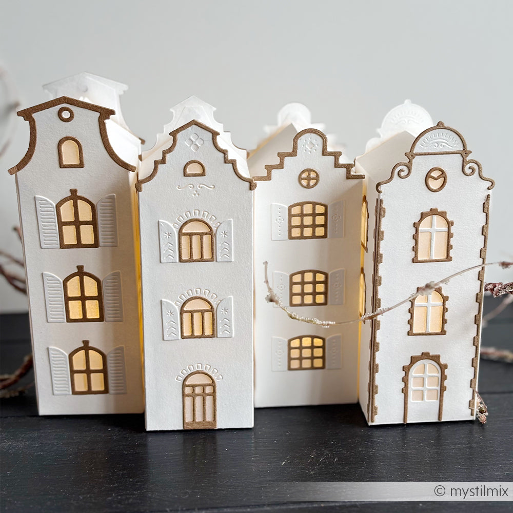 Drei weiße Papierlaternen in Form von dekorativen Häusern, gefertigt aus Designpapieren und mit der Motivstanze „Häuserbox 4“ von DieCut Systeme, leuchten durch ihre Fenster, während sie auf einer dunklen Unterlage stehen.
