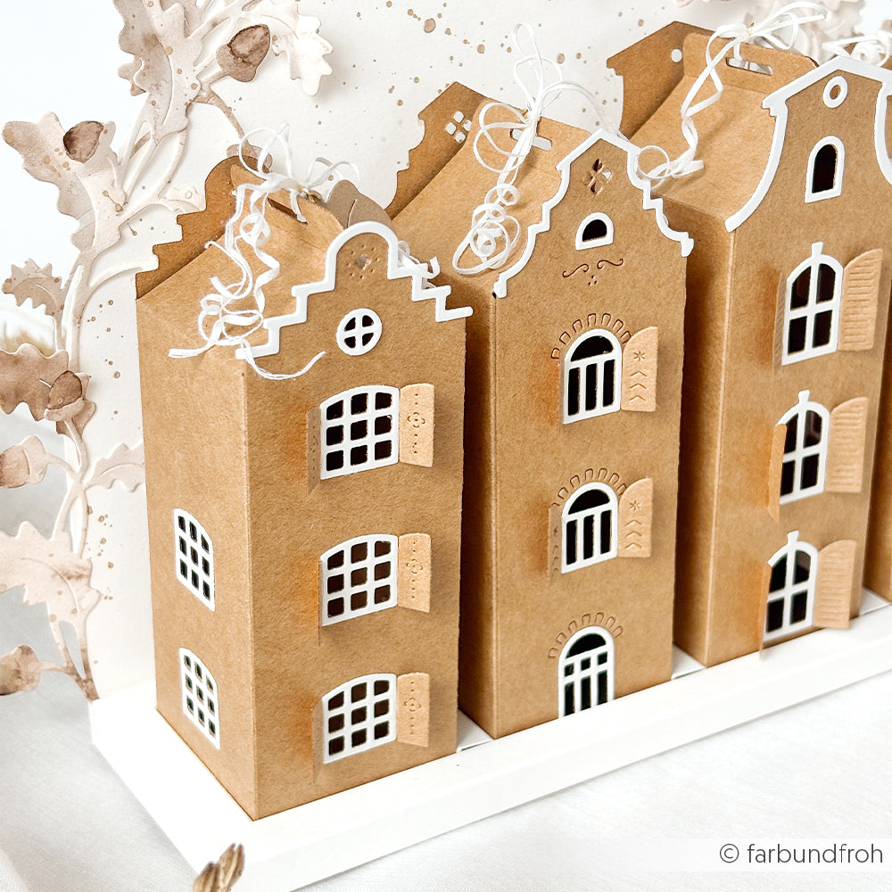 Drei braune Papierhäuschen mit weißen Fenster- und Türdetails, verziert mit weißen, geschwungenen Bändern. Diese bezaubernden Dekorationen, die mit der Motivstanze „Häuserbox 4“ und hochwertigen Designpapieren gestaltet wurden, unterstreichen die Vielseitigkeit moderner Stanzsysteme.