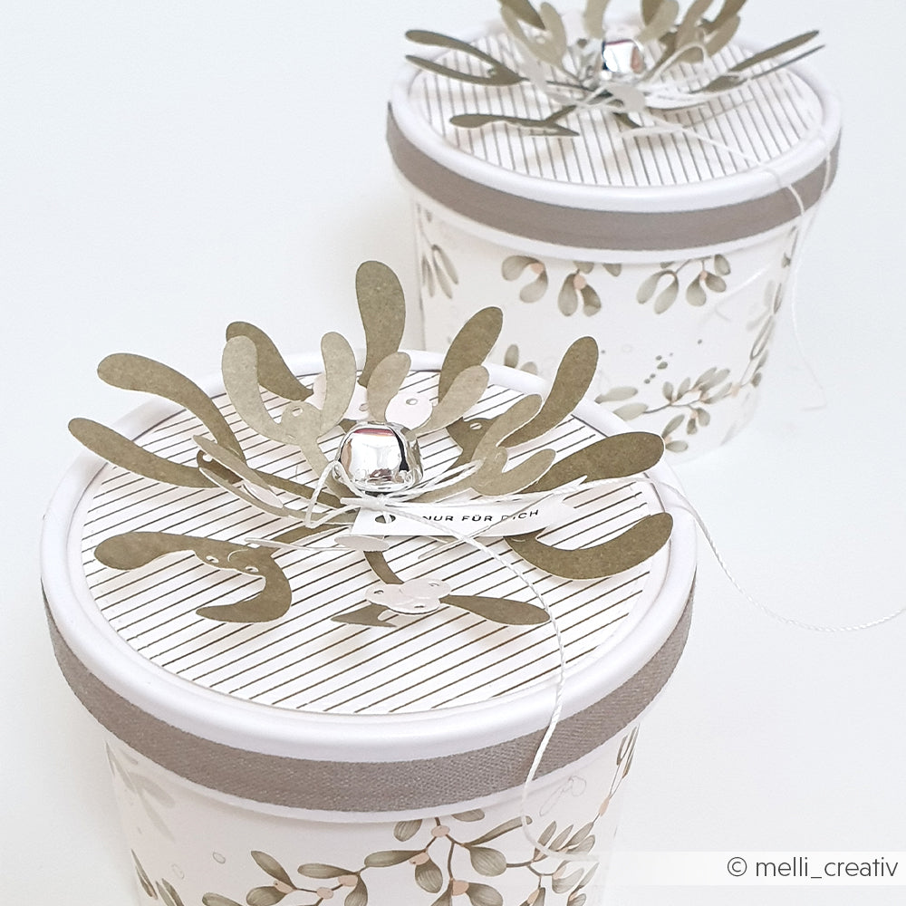 Zwei runde Geschenkboxen, verziert mit 3D-Ausstechmotiven in Form von Mistelzweigen und einem kleinen silbernen Glöckchen.