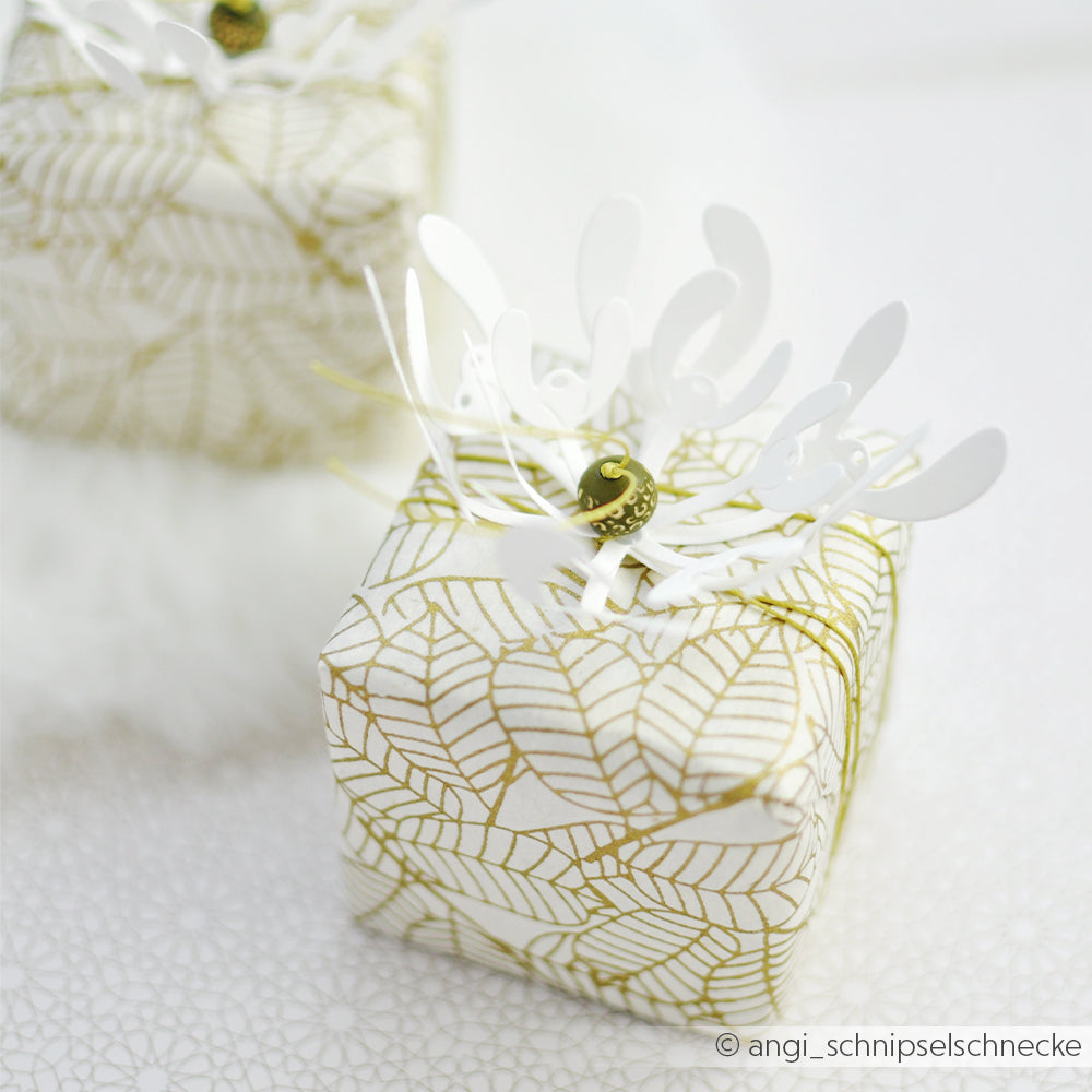 Zwei kleine Geschenkboxen, verpackt in Designpapier mit Blattgoldmuster und verziert mit dreidimensionalen Mistelzweigen als Blumenverzierung.