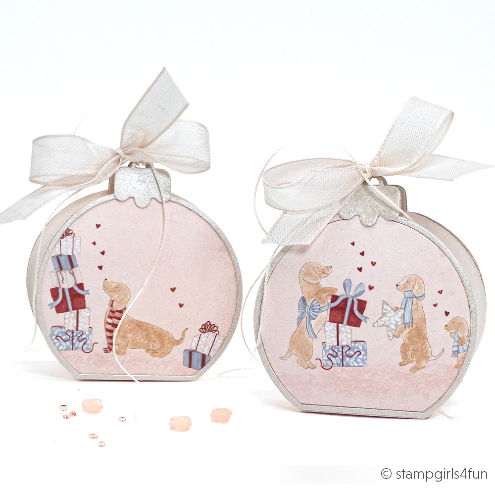 Zwei runde Geschenkboxen, verziert mit Dackeln und verpackten Geschenken, gekrönt von cremefarbenen Schleifen und verziert mit einem Motivstanze-Weihnachtskugel-Motiv.