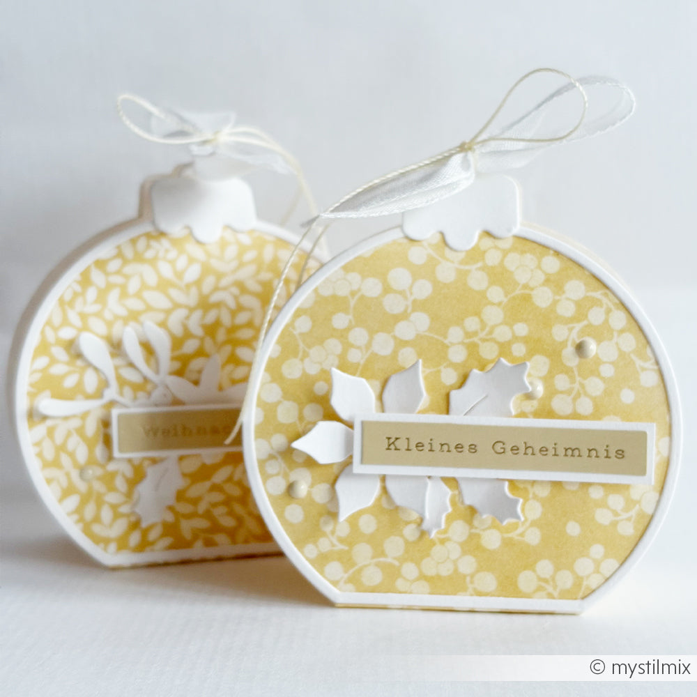 Zwei runde, gelbe, schmuckartige Geschenkboxen, die mit einer Motivstanze „Weihnachtskugel-Box“ hergestellt wurden und auf deren Vorderseite jeweils die Aufschrift „Kleines Geheimnis“ zu sehen ist.
