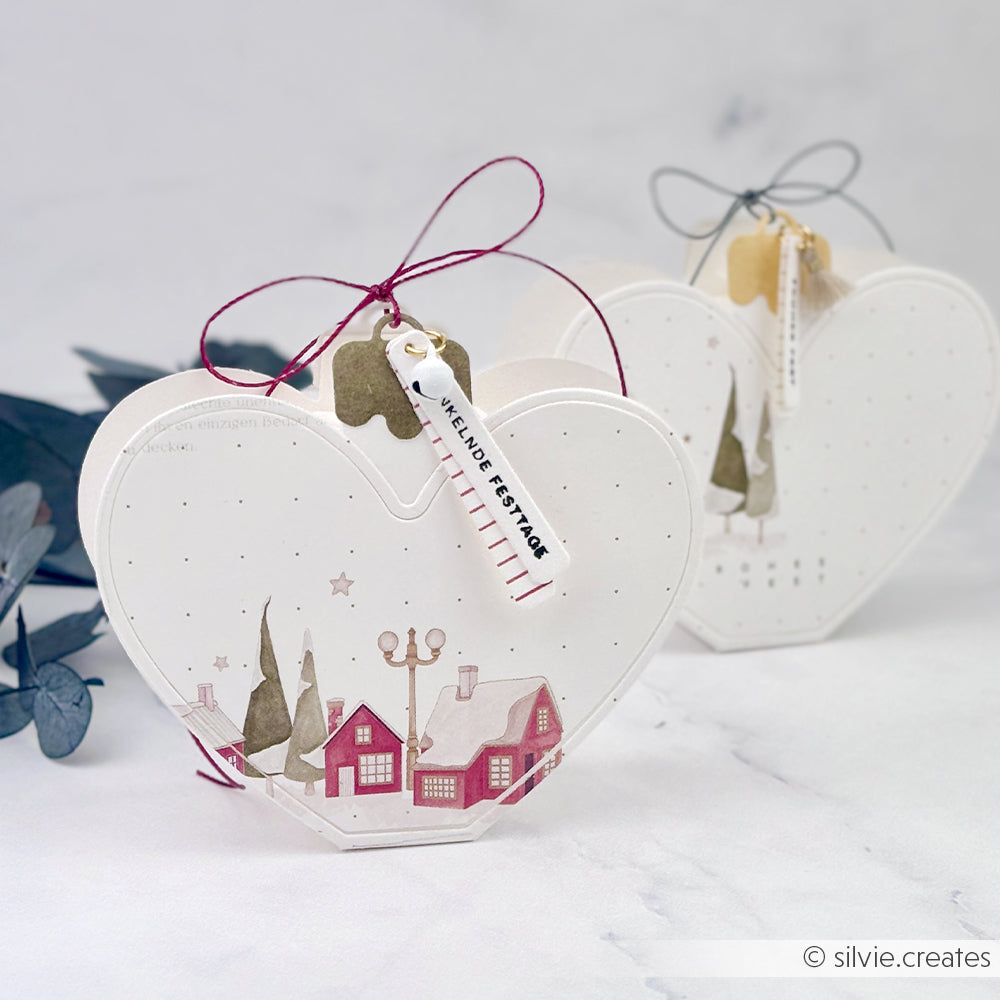 Zwei herzförmige Motivstanze-Geschenkboxen, verziert mit Motiven eines Winterdorfes und Anhängern.
