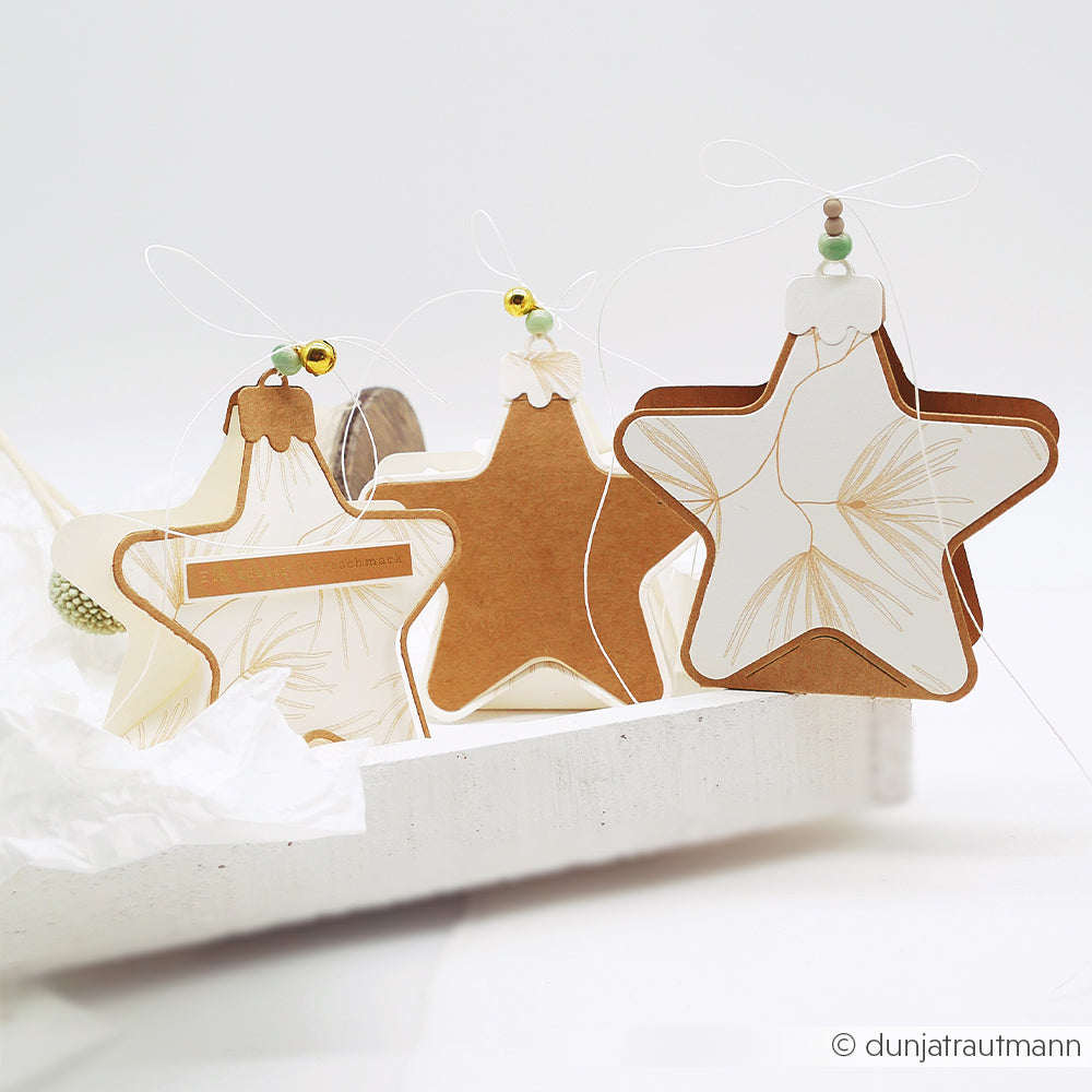 Drei sternförmige, ausgestanzte Weihnachtskugeln mit Tannennadel-Motiven, die an Schnüren hängen.