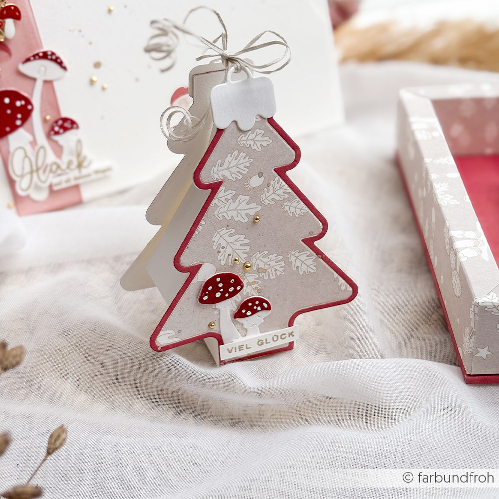 Eine Geschenkbox in Form eines Weihnachtsbaums, gefertigt aus Designpapier und verziert mit weißen Blattmustern und roten Pilzillustrationen.