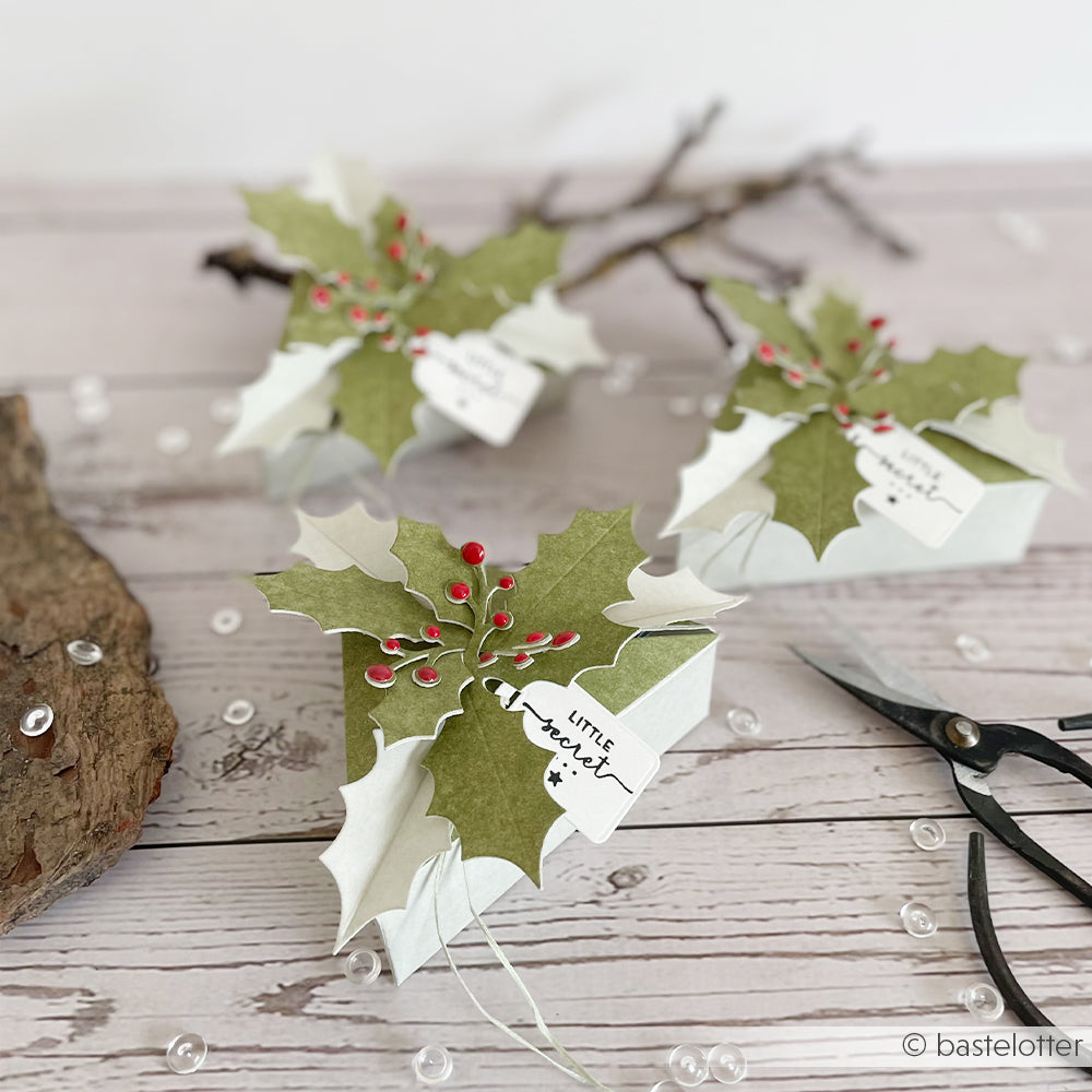 Drei dreieckige Ilex Box-Geschenkboxen, verziert mit Papier-Stechpalmenzweigen, Beeren und geheimnisvollen Anhängern.