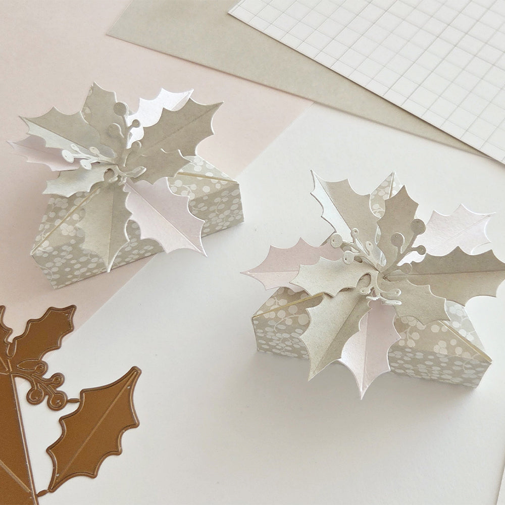 Zwei kleine, gemusterte Geschenkboxen, verziert mit Stechpalmenblättern aus Designpapier.
