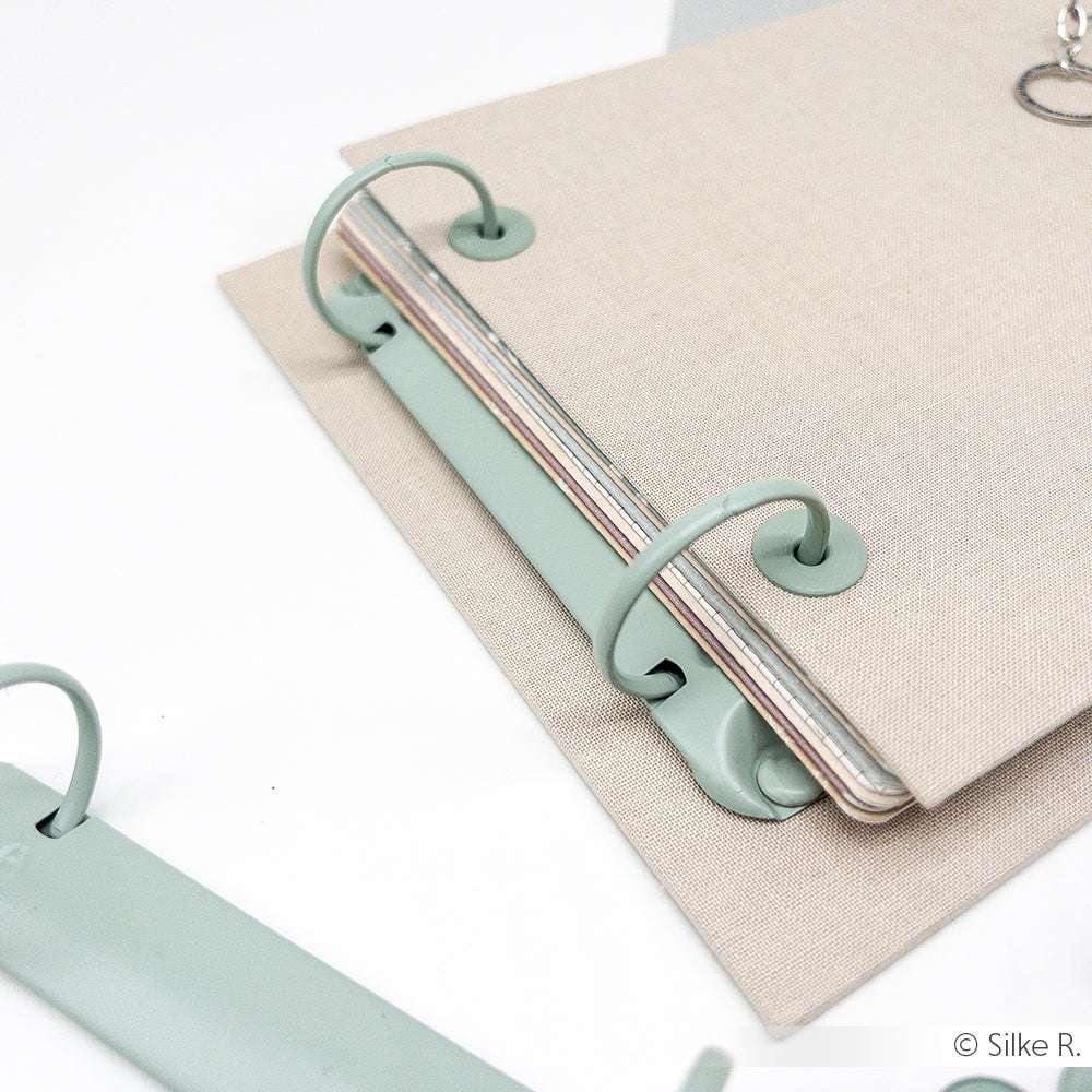 Der beigefarbene Alexandra Renke Binder mit grüner Ringmechanik fasst pastellfarbene Seiten, ideal für Mini-Alben oder Buchbindeprojekte, und ruht auf einer weißen Oberfläche.