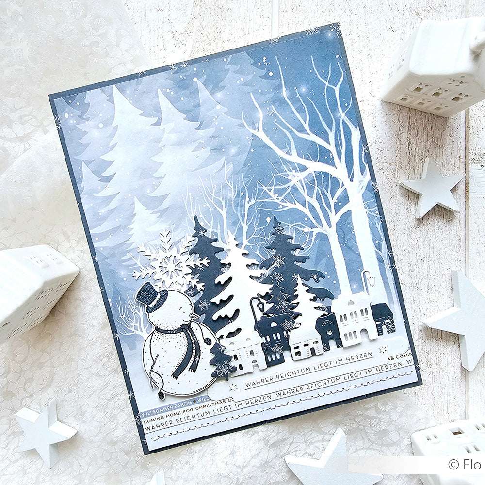Handgefertigte Winterkarte von Alexandra Renke mit einem Schneemann, Bäumen und Häusern in einem verschneiten blau-weißen Wald - hergestellt mit hochwertigem Designpapier für Ihre Papierprojekte.