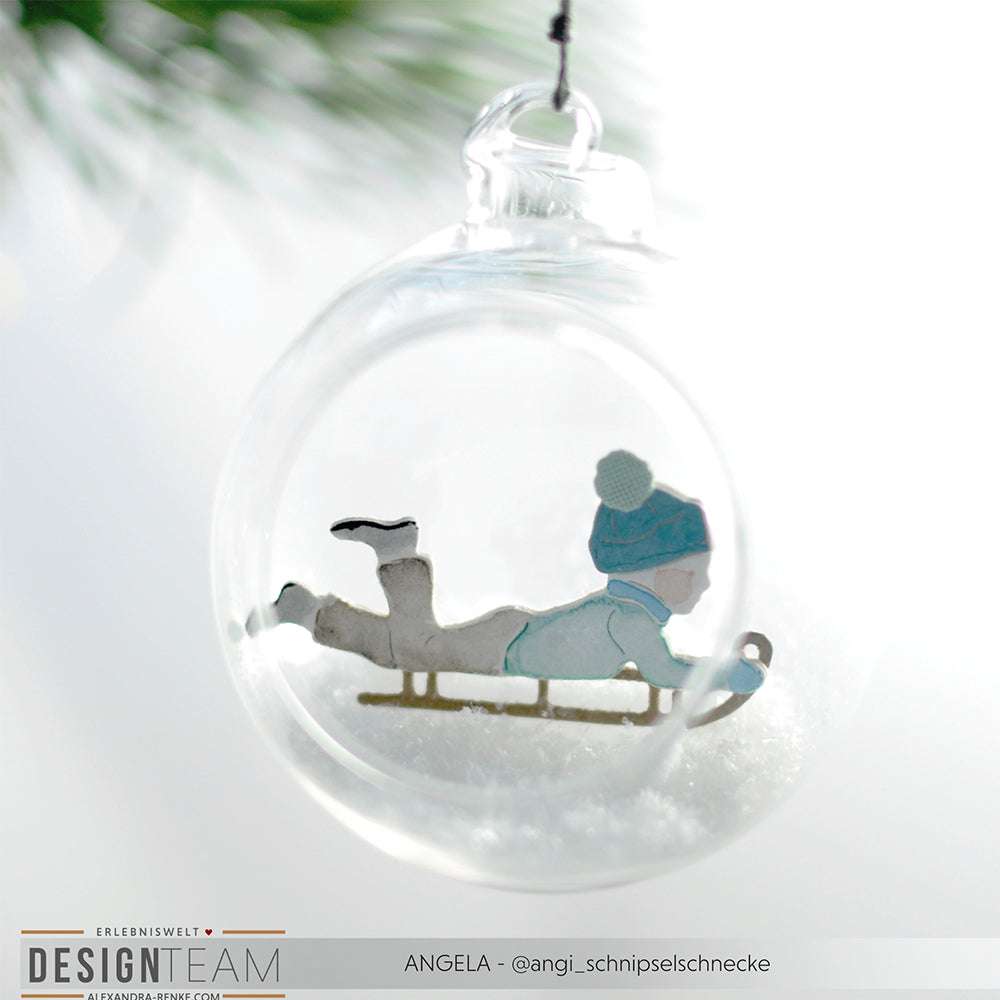 Klarglas-Ornament von Alexandra Renke mit einer Schlittenkinder-Figur, die mit einer blauen Mütze und Winterkleidung Schlitten fährt - ein ideales Weihnachtsdekor für Fans von einzigartigen Designpapieren.