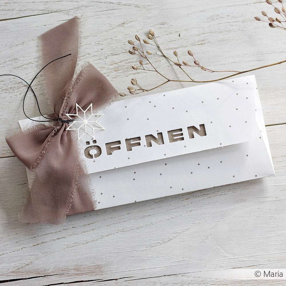 Weißer Briefumschlag von Alexandra Renke aus Designerpapier mit ausgestanztem "ÖFFNEN", gebunden mit einer lilafarbenen Schleife und einem Sternanhänger, ausgestellt auf einer hellen Holzfläche.