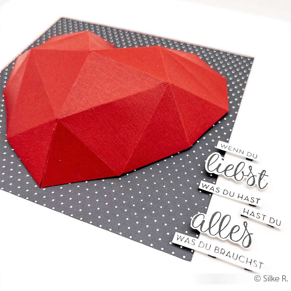 Ein rotes geometrisches Papierherz auf grauem Tupfenhintergrund mit deutschem Liebestext, hergestellt aus 100% recyceltem Feinleinen Cardstock von ☆ für Eleganz und Nachhaltigkeit.