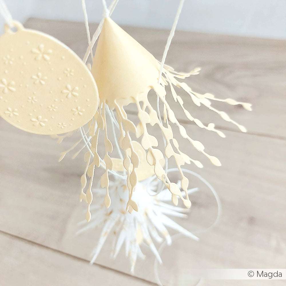 Zarte Papierornamente aus floralen Designpapieren von Alexandra Renke, mit ausgeschnittenen Fransen, die mit Motivstanze Dies hergestellt wurden, hängen an Schnüren vor einem hellen Holzhintergrund.