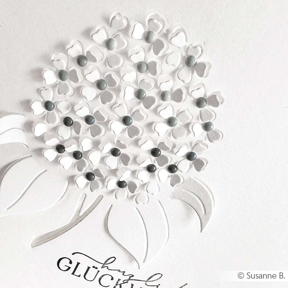 Weißes Strukturpapier einer Hortensienblüte mit erhabenen Blütenblättern und kosmosblauen Emaille-Punkten, darunter der Text "Glück" - eine elegante Kartengestaltung von Alexandra Renke.