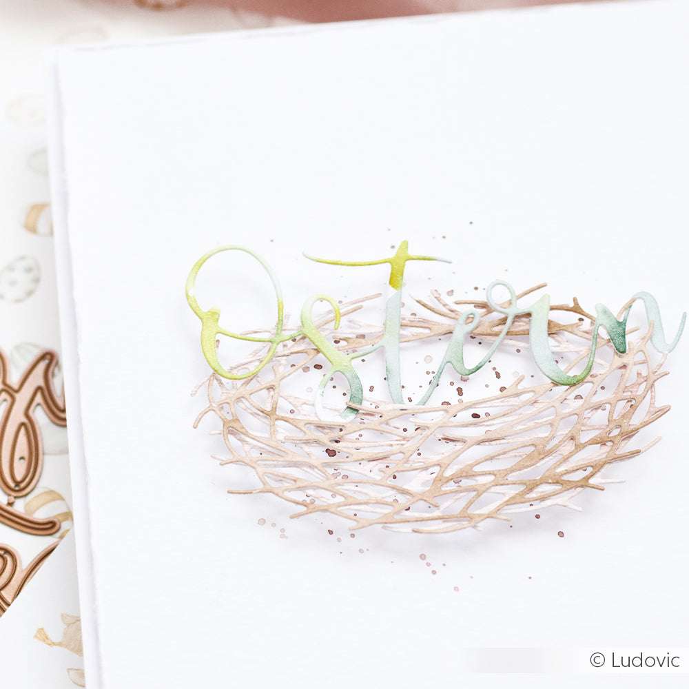 Eine weiße Karte ziert ein Papiernest in Pastelltönen mit dem Schriftzug "Frohe Ostern", gestaltet mit einer Motivstanze von Alexandra Renke.