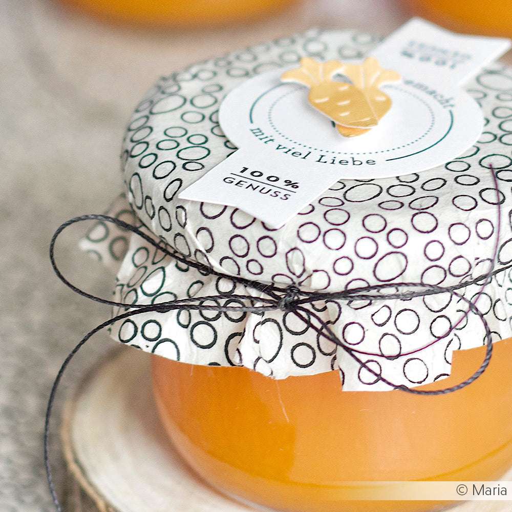 Ein Glas mit Orangenmarmelade von Alexandra Renke, mit gemustertem Deckel, Makramee-Schnur und der Aufschrift "100% Genuss und viel Liebe".