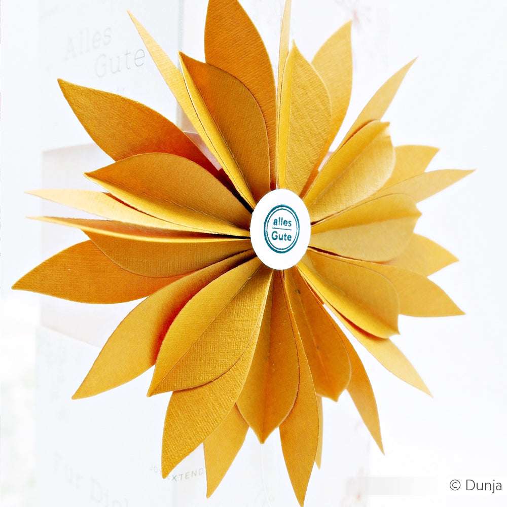Eine goldene Papierblume von ☆ aus recyclebar Spezialpapier mit einem runden "Alles Gute"-Anhänger in der Mitte schmückt ein weißes Geschenk.