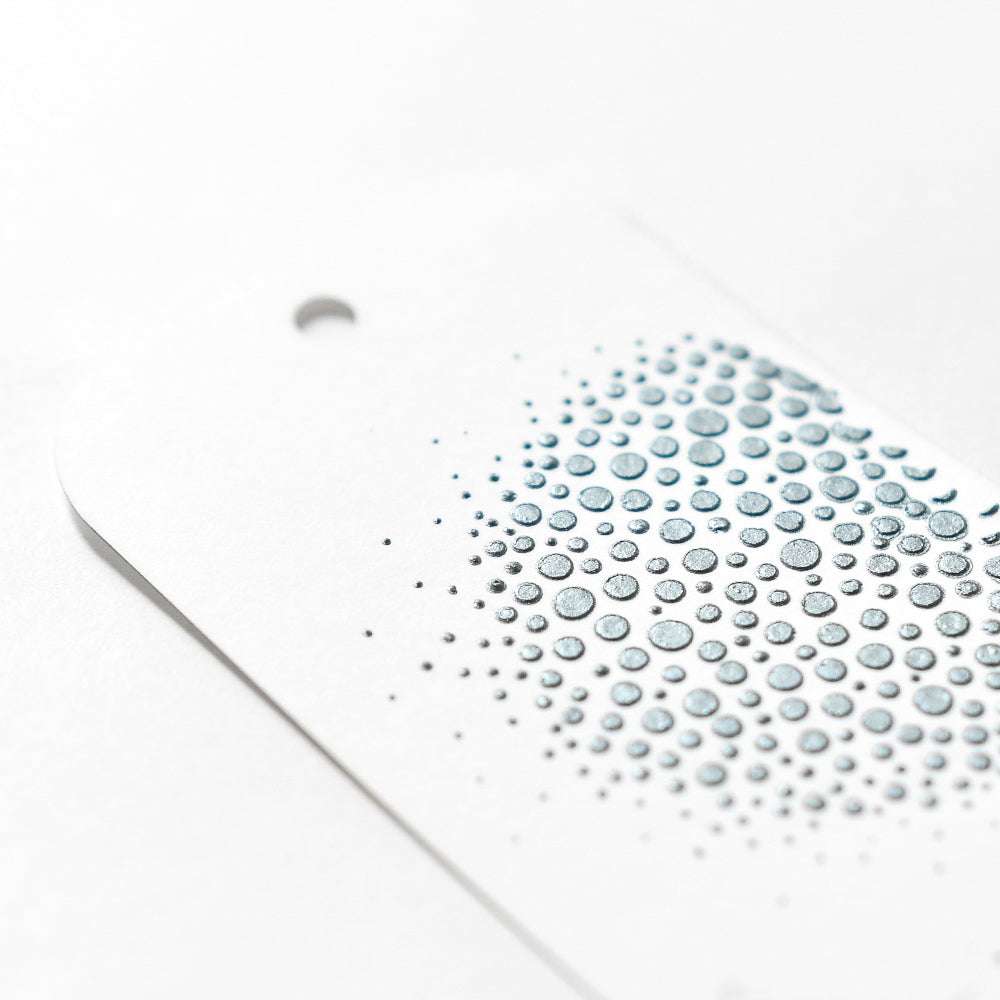 Ein weißer Anhänger von The Crafters Workshop mit geprägten silbernen Punkten in einem Farbverlauf, hergestellt mit Schablonen und Modellierpaste, und einem Loch oben links.