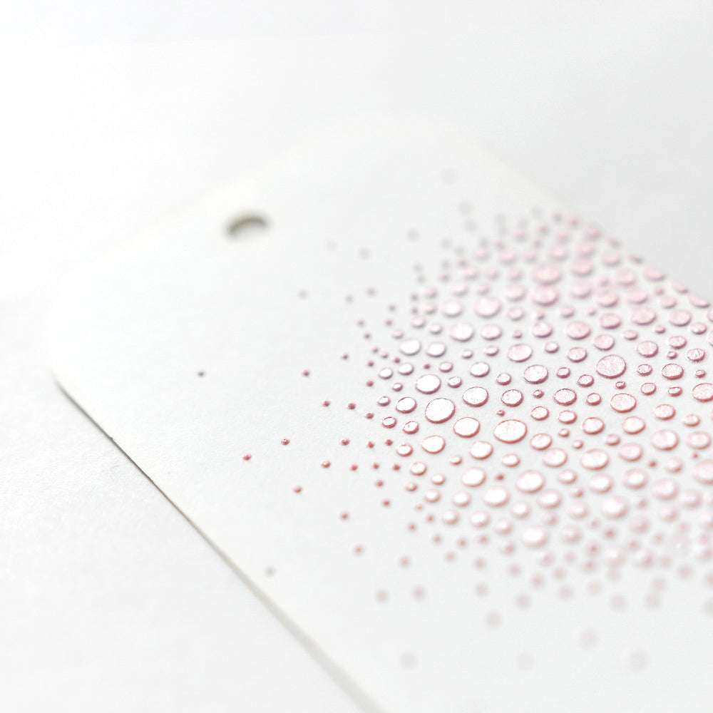 Ein weißer Anhänger von The Crafters Workshop mit einem Loch und erhabenen rosafarbenen Punkten in einem Farbverlaufsmuster; das Design schimmert - ideal, um porösen Oberflächen eine Struktur zu verleihen oder um es mit Stencil Butter zu kombinieren.