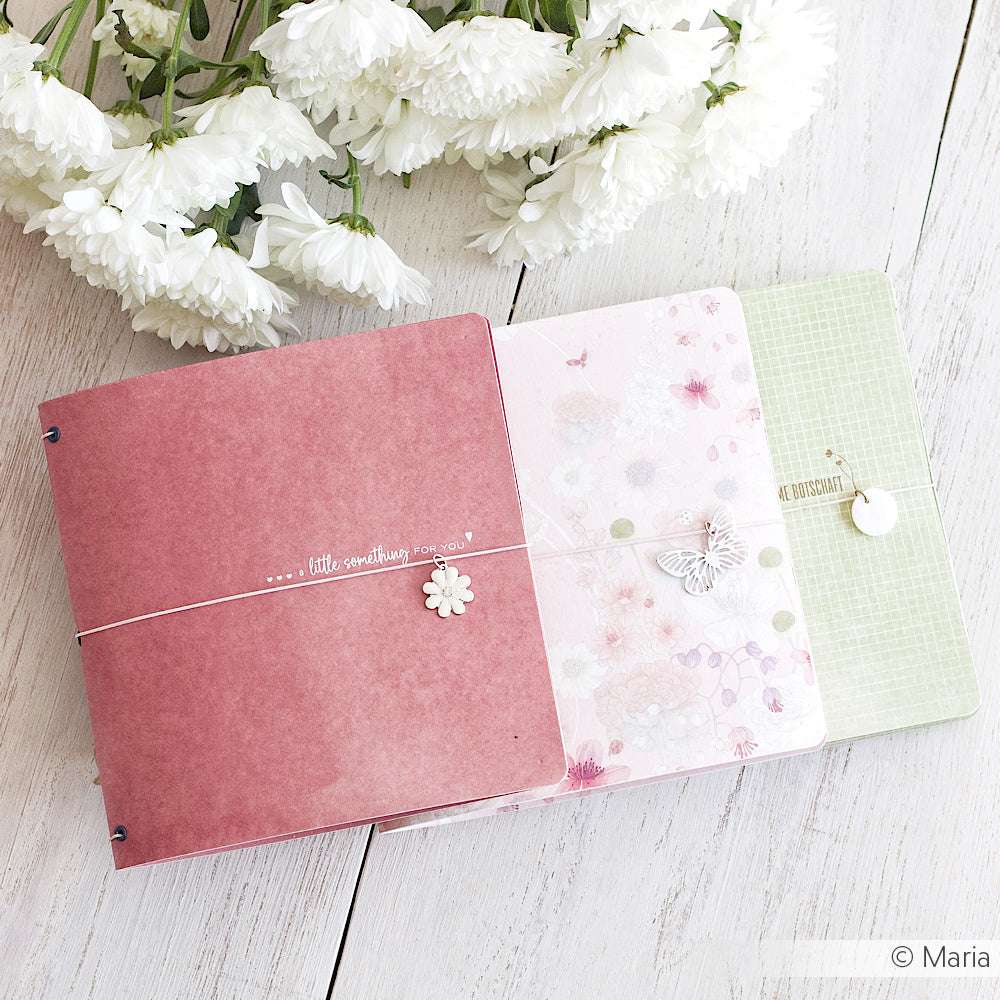 Drei pastellfarbene Zeitschriften mit floralen Motiven von Alexandra Renke, gefertigt aus edlem Designpapier, sind auf einer weißen Holzfläche angeordnet und mit zarten weißen Blüten darüber akzentuiert.