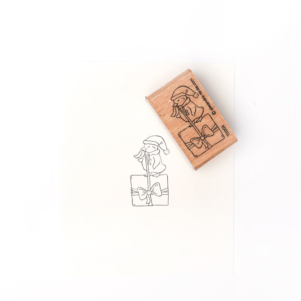 Ein Alexandra Renke Holzstempel und sein Tuscheabdruck zeigen einen Wichtel mit Weihnachtsmannmütze, der auf einer eingepackten Geschenkschachtel sitzt, mit detailliertem Stempelgummi - perfekt für festliche Bastelarbeiten.