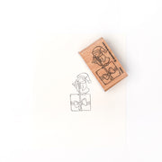 Ein Alexandra Renke Holzstempel und sein Tuscheabdruck zeigen einen Wichtel mit Weihnachtsmannmütze, der auf einer eingepackten Geschenkschachtel sitzt, mit detailliertem Stempelgummi - perfekt für festliche Bastelarbeiten.