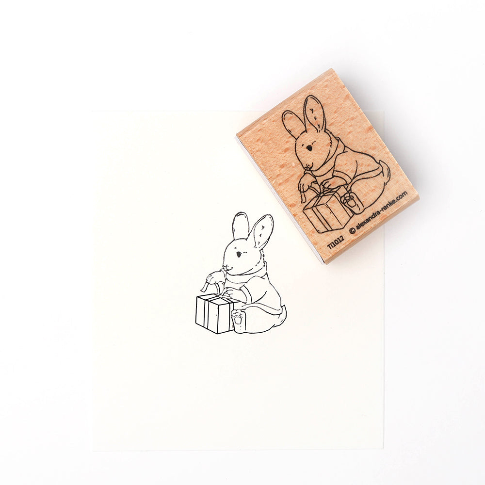 Ein Alexandra Renke Holzstempel und der dazugehörige Druck zeigen einen Stempel Hase - ein Kaninchen in einem Mantel, das eine eingepackte Geschenkschachtel hält und betrachtet.