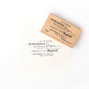 Ein Alexandra Renke Holzstempel druckt "Verpackt von Rudolf" in dekorativer deutscher Schrift auf weißem Papier.
