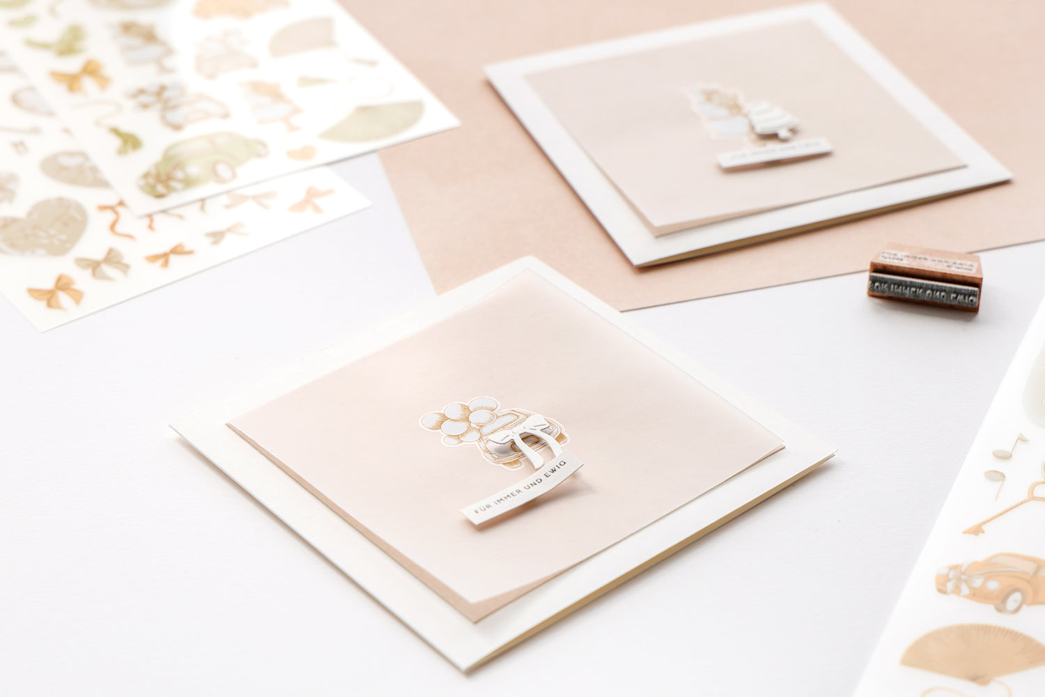 Zwei handgefertigte quadratische Grußkarten in sanften Beige- und Weißtönen, dekoriert mit Rub-On-Stickern eines Hochzeitsautos mit Luftballons und dem Schriftzug 'Für immer und ewig', umgeben von weiteren Bastelutensilien auf einem weißen Untergrund.
