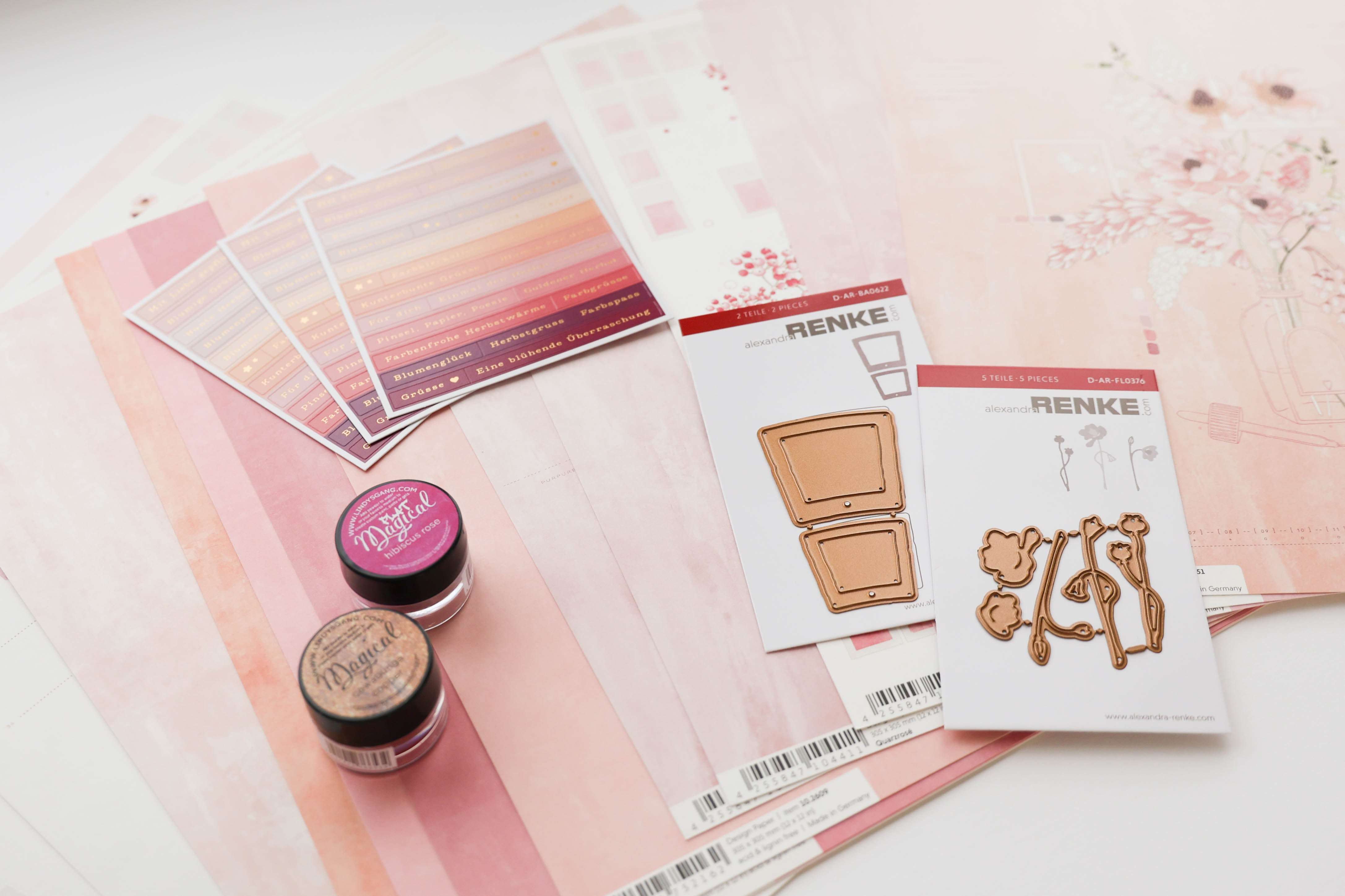 Bastelbedarf in Rosa, darunter Papier, Aufkleber, Tintentöpfe und Stanzformen aus Metall, angeordnet auf einer weißen Fläche.