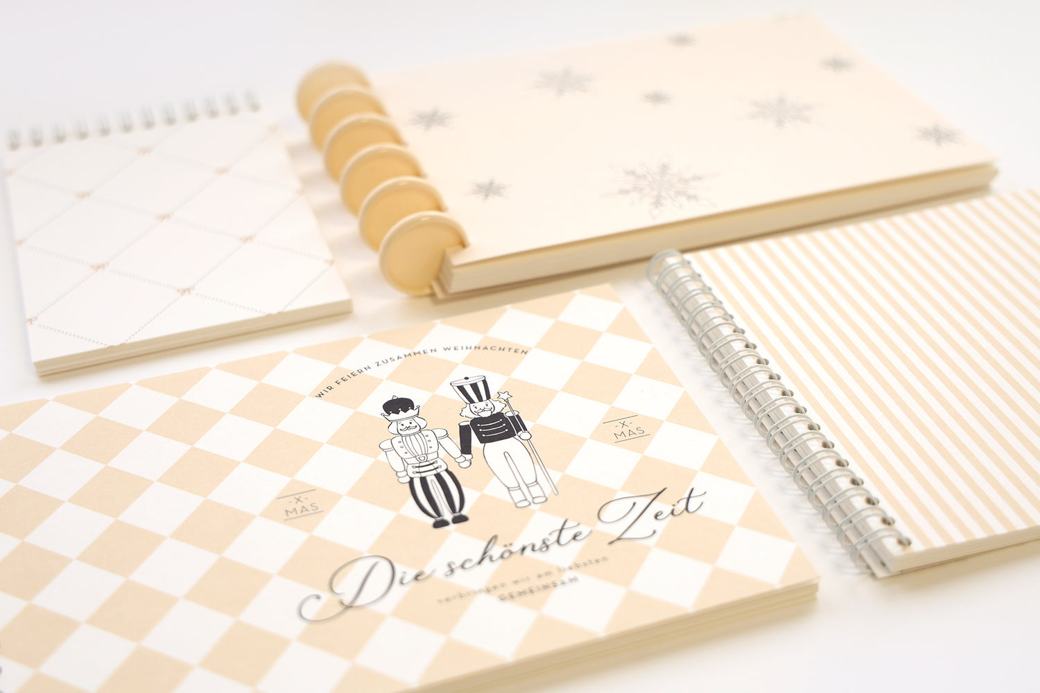 Vier beigefarbene und weiße Notizbücher mit Mustern und deutschem Text, ordentlich auf einer weißen Fläche angeordnet.