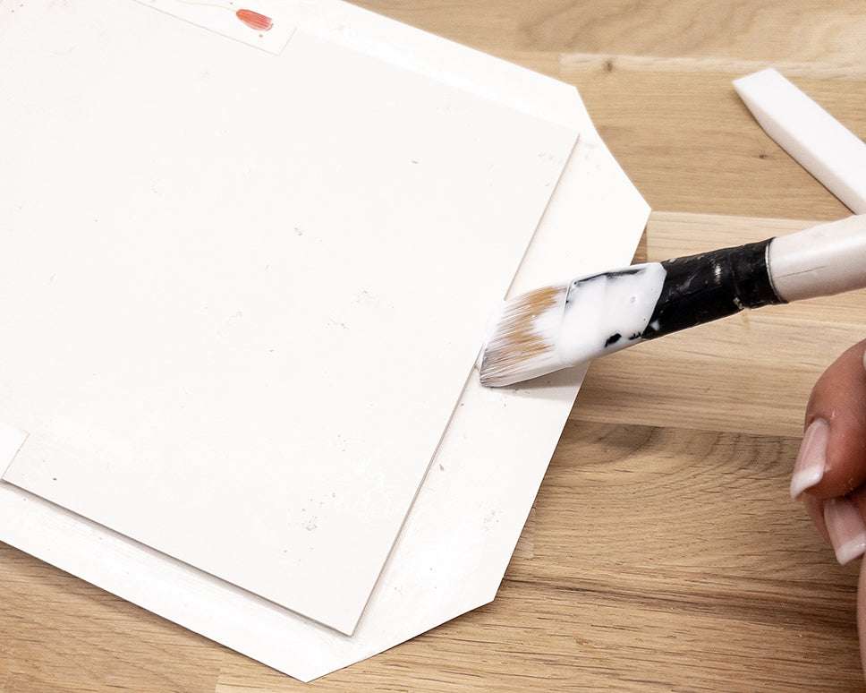 Eine Hand trägt mit einem Pinsel auf einer hölzernen Unterlage Leim auf den Rand eines weißen Papiers auf.