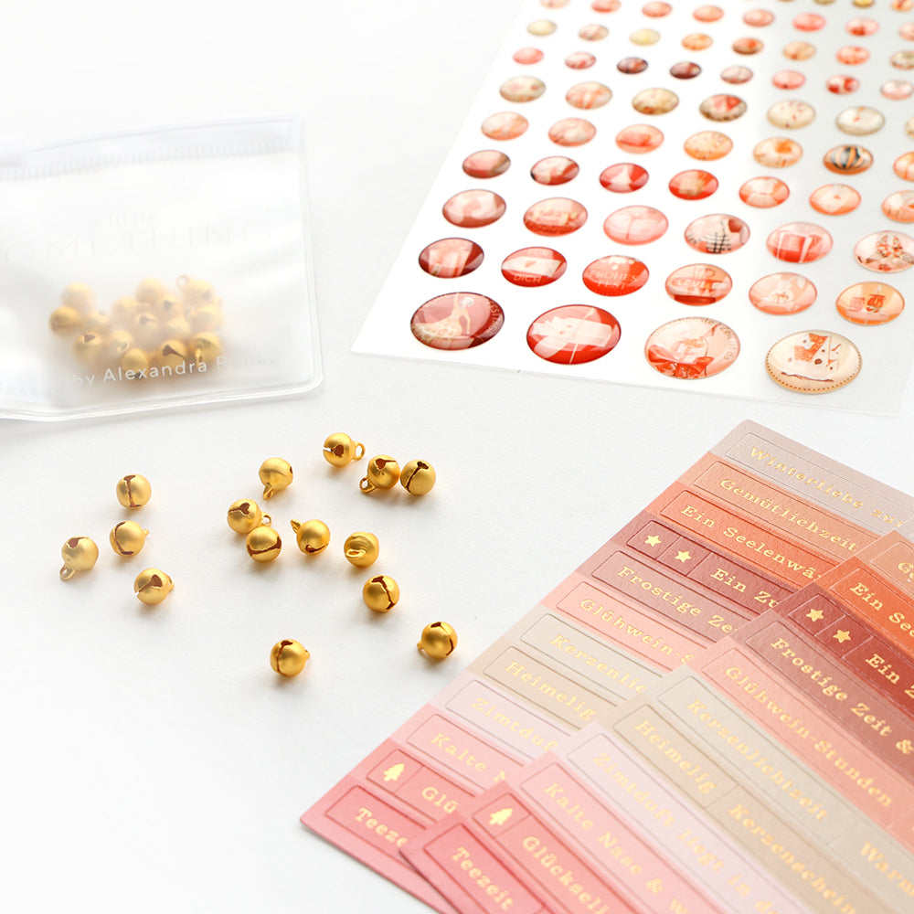 Goldene Jingle Bells, rote und rosafarbene Aufkleber und ein Beutel auf einer weißen Fläche.