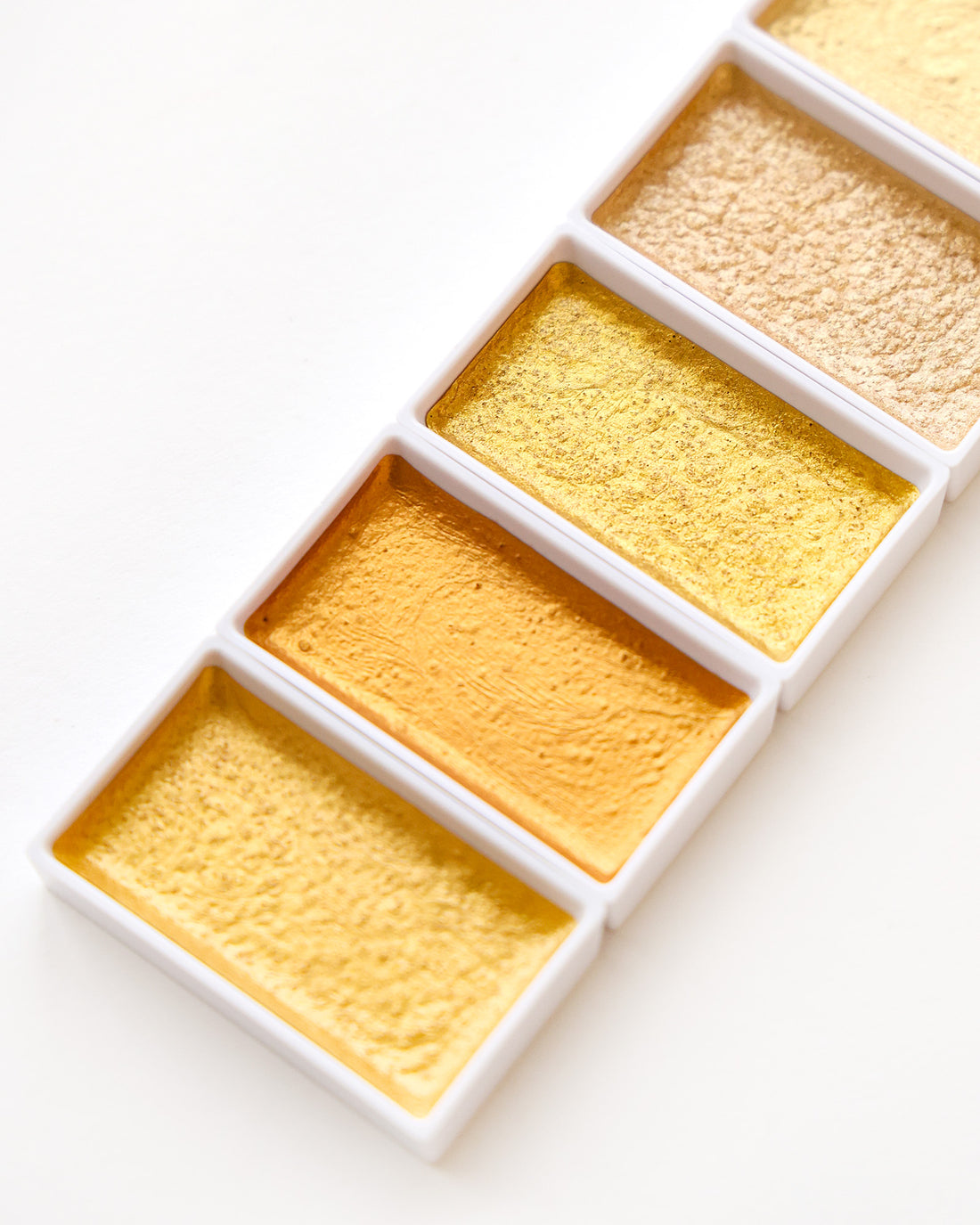 Vier quadratische Töpfe mit gelber und goldener Aquarellfarbe, die diagonal auf einem weißen Hintergrund angeordnet sind.