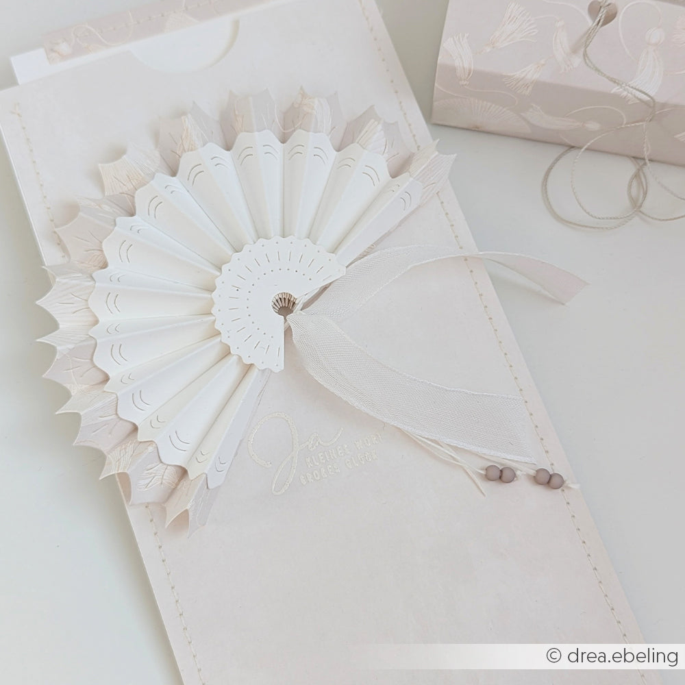 Eleganter weißer Papierfächer von Alexandra Renke, gefertigt aus Designpapieren und veredelt mit Schleife und Perlen auf strukturiertem Karton. Zarte Details und geprägter Text, hergestellt mit der Motivstanze Faltfächer DieCut Systeme.