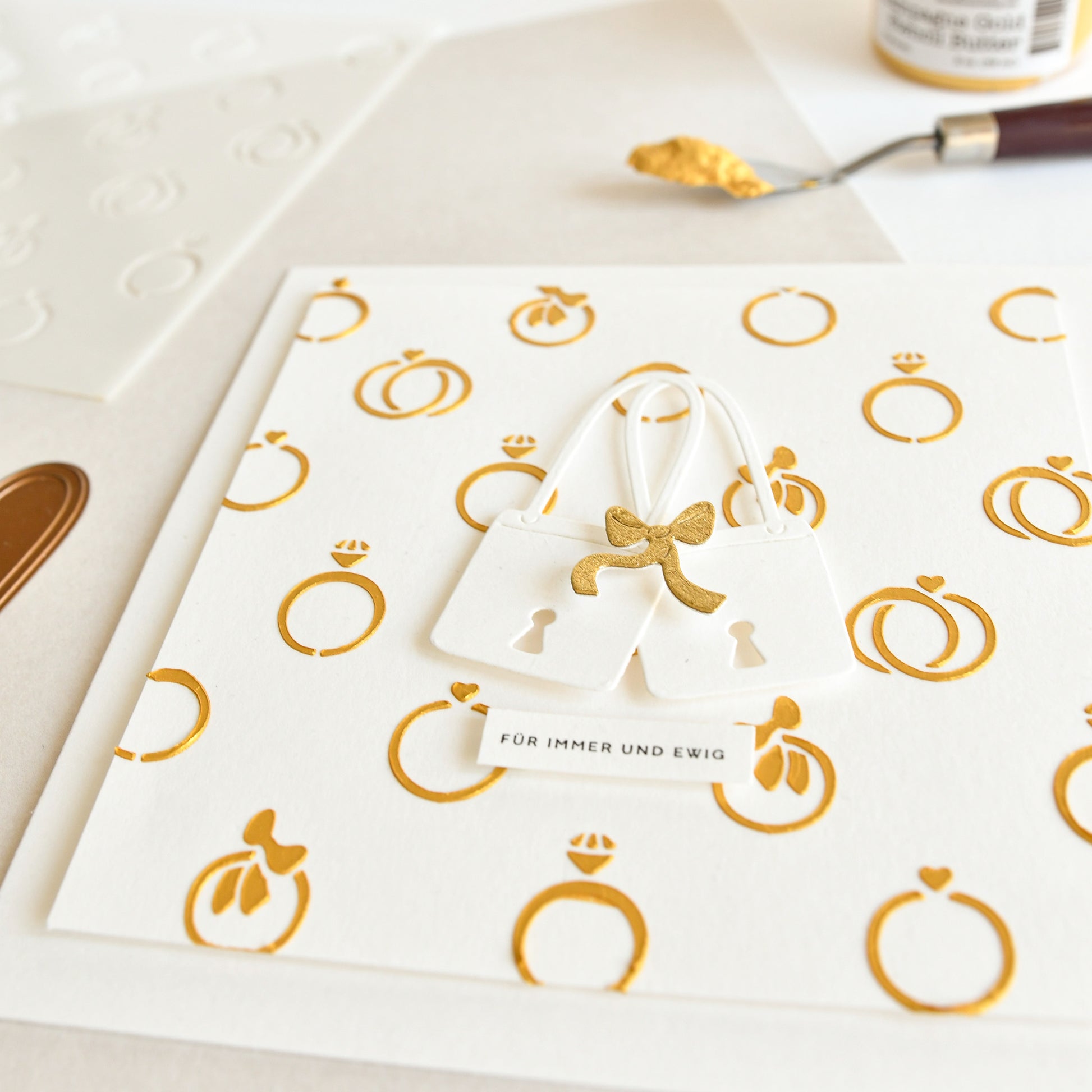 Hochzeitskarte von Alexandra Renke mit elegantem Designpapier, goldenen Ringmustern, zwei weißen Schlossanhängern, die mit einer Zierschloss-Stanze hergestellt wurden, und einem deutschen Etikett: "Für immer und ewig".