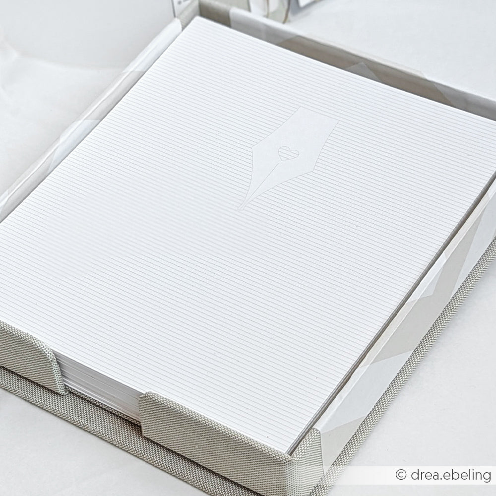 Ein Stapel weißes, gefüttertes Briefpapier mit geprägter Schreibfeder liegt in einer stoffbezogenen Zettelbox von Alexandra Renke, die aus hochwertigen Graupappe-Zuschnitten für lange Haltbarkeit gefertigt ist.
