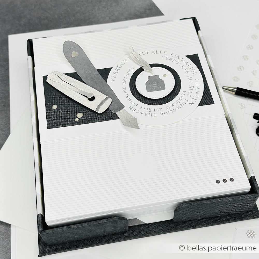 Eine handgefertigte monochrome Karte mit geschichteten Papierwerkzeugen und deutschem Text, hergestellt mit Alexandra Renke PAPPKit Graupappe Zuschnitte, präsentiert auf einem schwarzen Tablett.