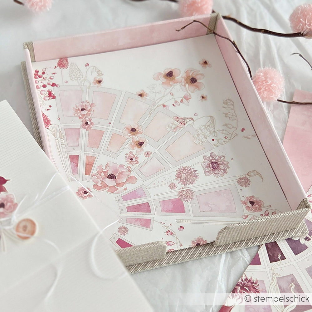 Ein dekoratives rosafarbenes Tablett mit Blumenmustern und Aquarellfarben von Alexandra Renke, daneben rosa-weißes Briefpapier und eine PAPPKit Zettelbox zur Organisation von Notizen.