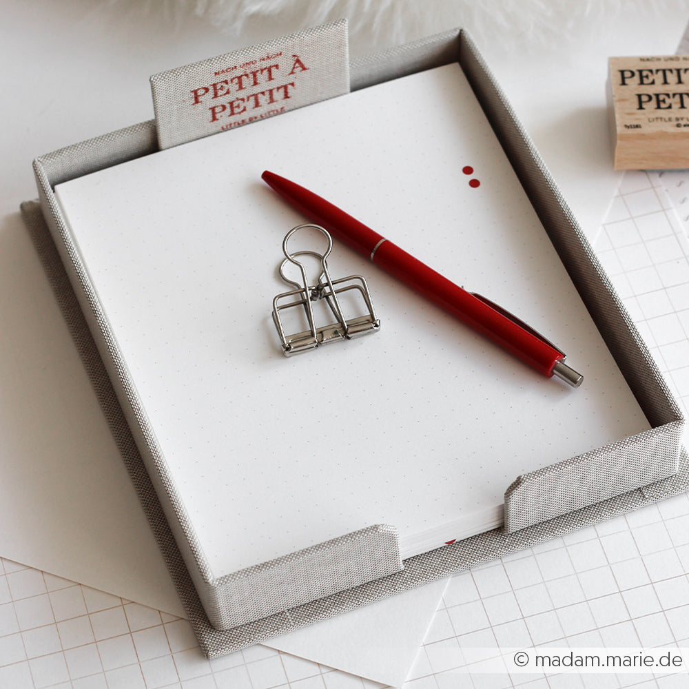 Ein roter Stift und eine Büroklammer liegen auf einem leeren Notizblock in einer Alexandra Renke Zettelbox aus Graupappe-Zuschnitten, die auf einem weißen Schreibtisch steht.