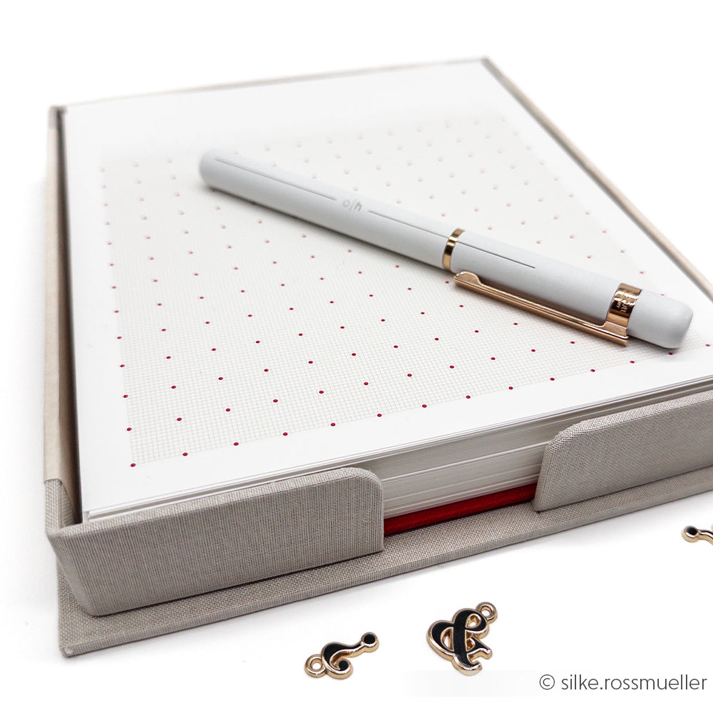 Ein weißer Stift liegt auf einem gepunkteten Notizblock in einem grauen Alexandra Renke Zettelbox-Halter, daneben befinden sich Metallzeichen.