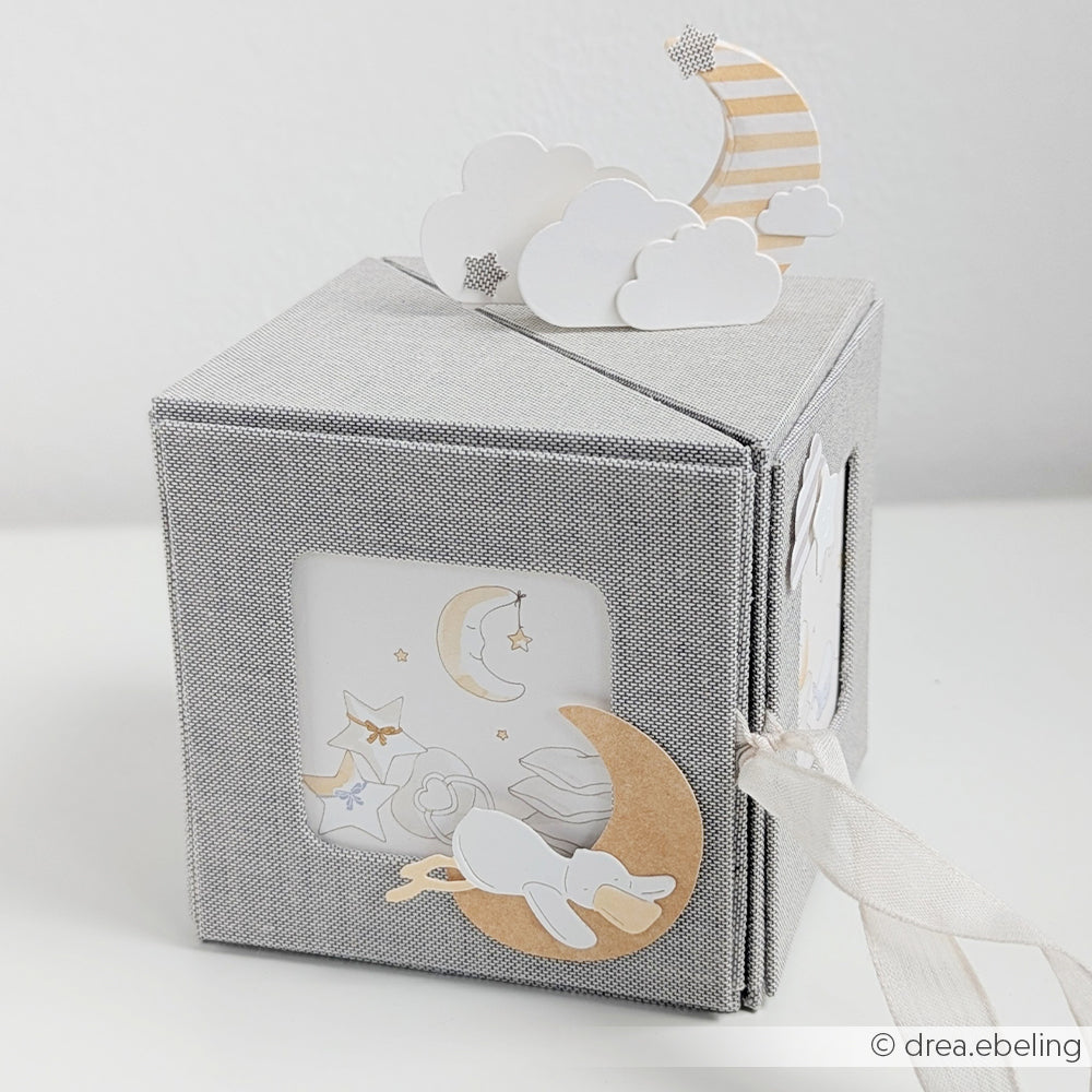 Fotowürfelbox aus grauem Stoff von Alexandra Renke, verziert mit Mond-, Wolken- und Sternenmotiven; ein Fenster offenbart einen schlafenden Hasen und himmlische Formen im Inneren.