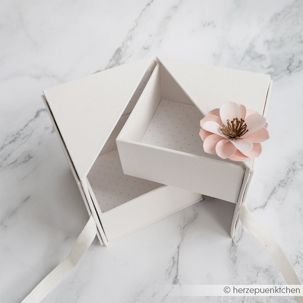 Alexandra Renke weiße dreieckige Fotowürfelbox mit einer rosa Papierblume, teilweise geöffnet und auf einer Marmorfläche ausgestellt.