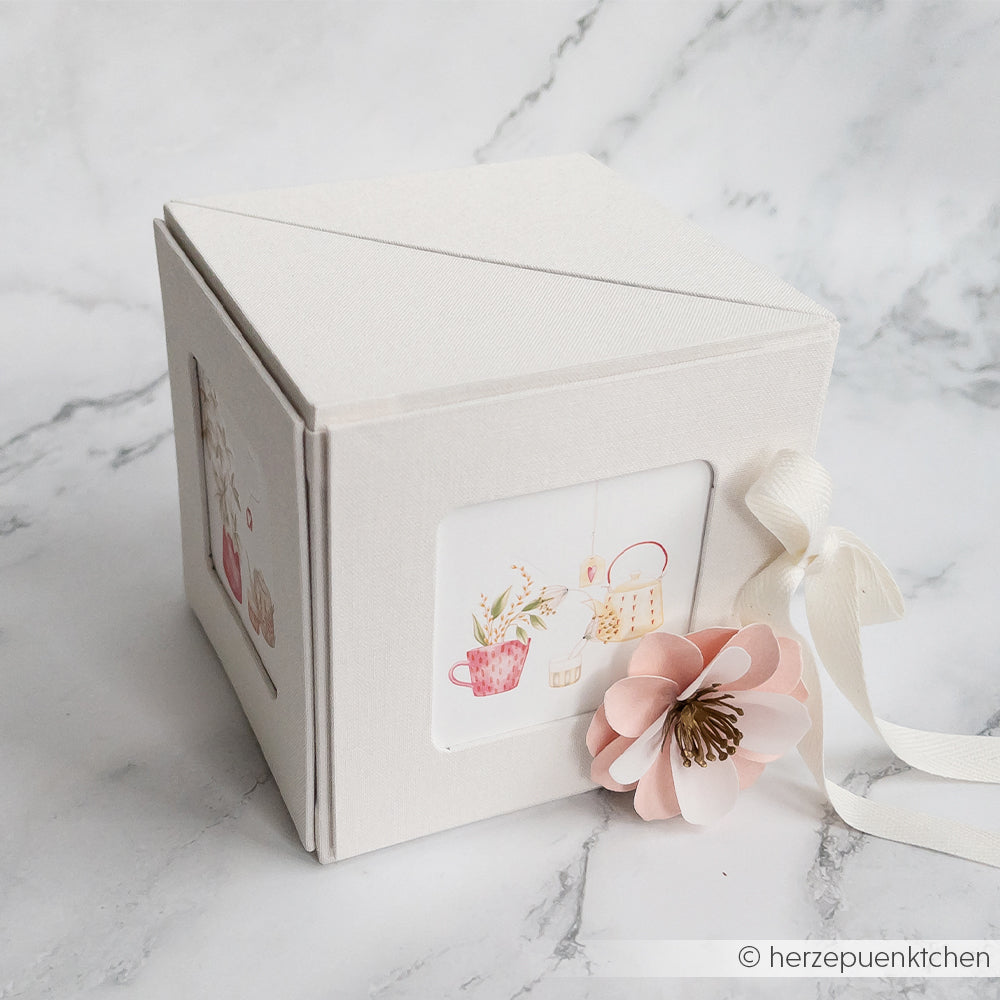 Die Alexandra Renke Fotowürfelbox ist eine dekorative weiße Geschenkbox mit einer rosa Papierblume und einer Illustration von Tassen und Pflanzen auf einer Seite.
