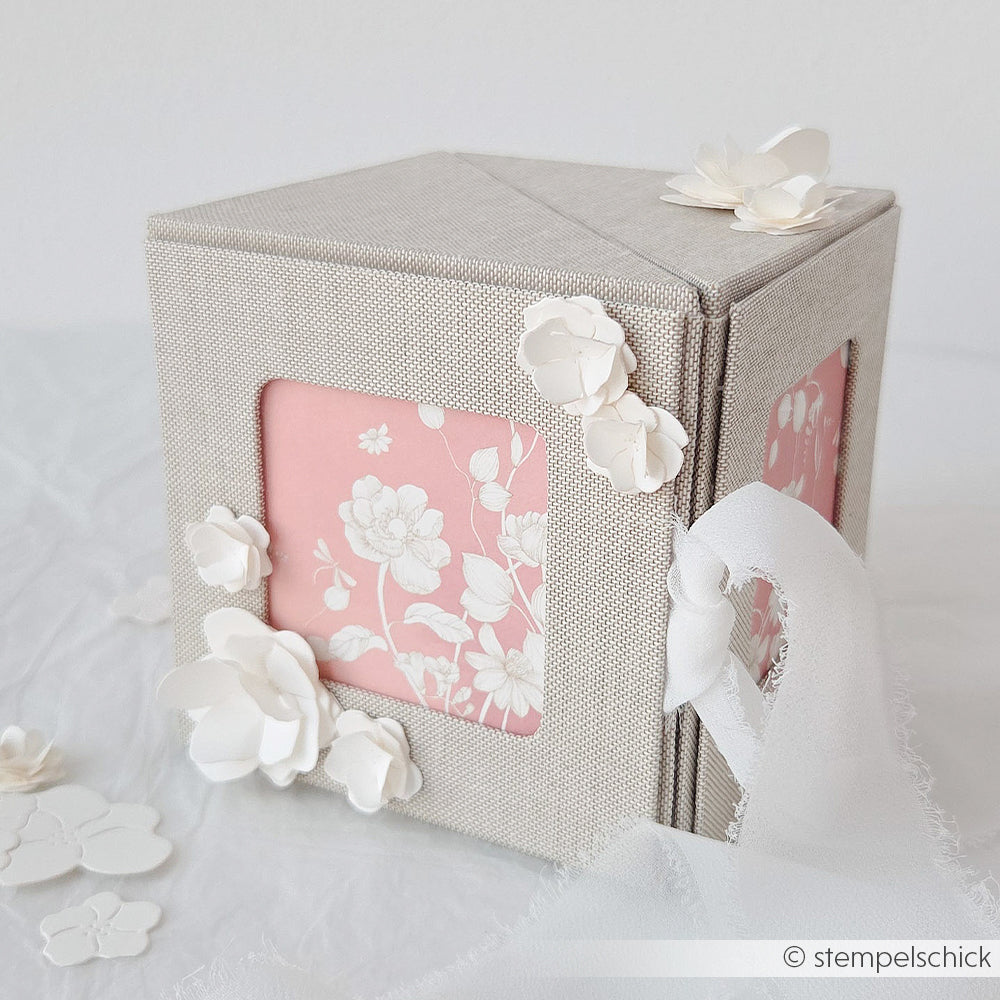 Eine dekorative grau-rosa Alexandra Renke Fotobox mit Blumenausschnitten, weißen Papierblumen und einem durchsichtigen Band.